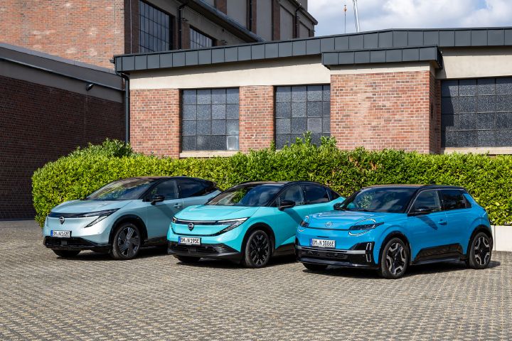 Vor einem Backsteingebäude und einer grünen Hecke stehen nebeneinander die drei Nissan EV-Modelle Nissan Ariya, Nissan Leaf und Nissan Micra, Blick von schräg vorne auf die Gruppe