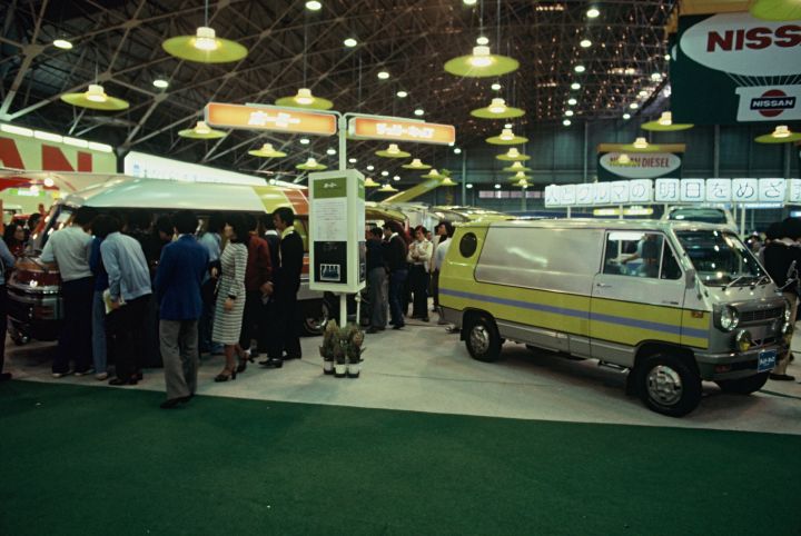 A bustling car exhibition displaying various vehicles, including a prominently featured white and yellow van. Attendees gather around exhibits under bright lights in a spacious venue with visible brand signage overhead.