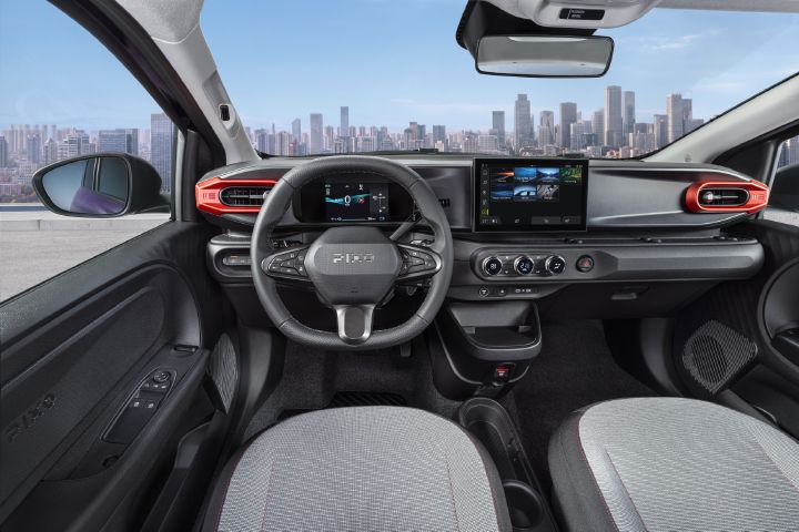 Photograph showcasing the all-new PIXO interior with steering wheel, digital instrument cluster, central touchscreen, climate controls, and textured seats. Red-accented air vents, black dashboard trim, compact center console, and city skyline visible through windshield emphasize vehicle design and urban driving context.