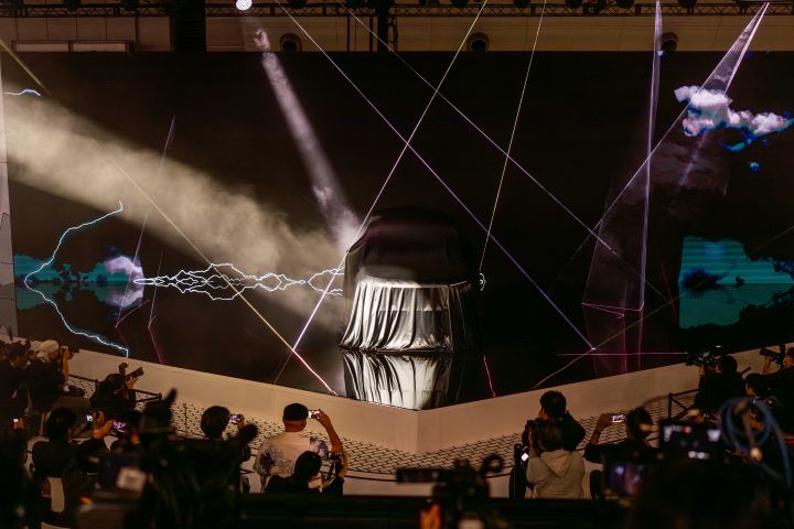 In a dark stage setting, the all-new Elgrand is positioned at the center, covered with a black cloth, while the background screen shows illuminated lines and visual effects. In the foreground, members of the press are gathered, taking photos with cameras.