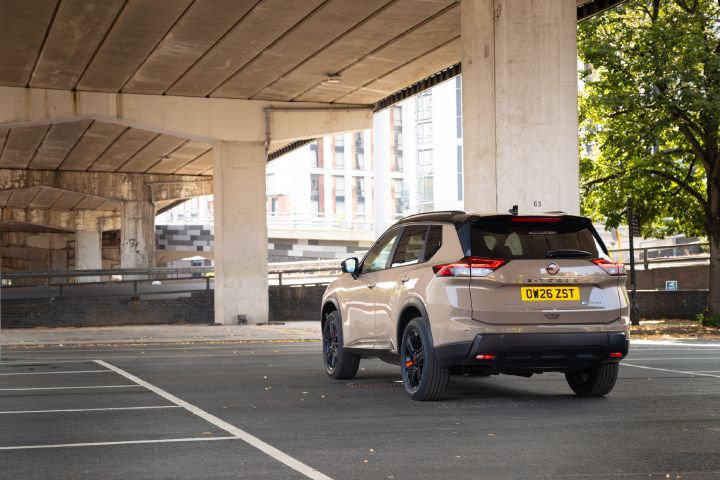 Rear view of a champagne-coloured Nissan X‑Trail parked in an urban setting, with trees, buildings and an overpass in the background