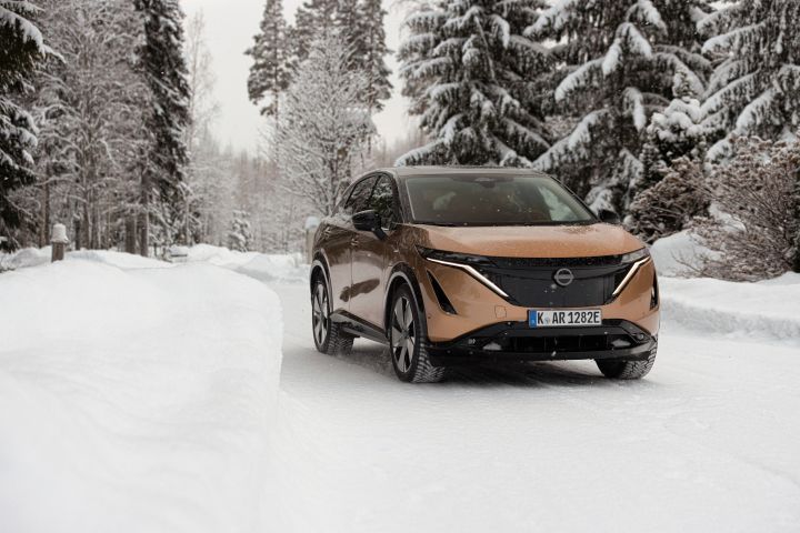 Elektroauto Nissan Ariya e‑4ORCE in Akatsuki Copper mit Dach und Außenspiegel in Pearl Black auf schneebedeckter Straße mit verschneiten Bäumen auf beiden Seiten, 3/4 Front