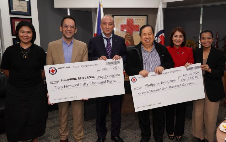 Six individuals stand in an office setting holding two oversized donation checks from Nissan Philippines, Inc. to the Philippine Red Cross. One check is for ₱250,000 and the other is for ₱19,150, both dated July 10, 2026. Framed certificates, flags, and office furnishings are visible in the background. The group poses behind the checks for a commemorative photo documenting the donation turnover.