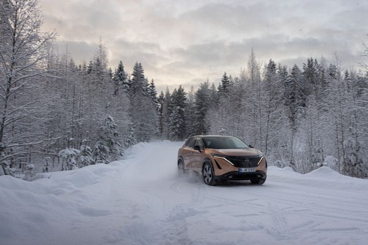 Elektroauto Nissan Ariya e‑4ORCE in Akatsuki Copper mit Dach und Außenspiegel in Pearl Black fährt im Sonnenuntergang auf schneebedeckter Straße mit verschneiten Bäumen auf beiden Seiten, 3/4 Front