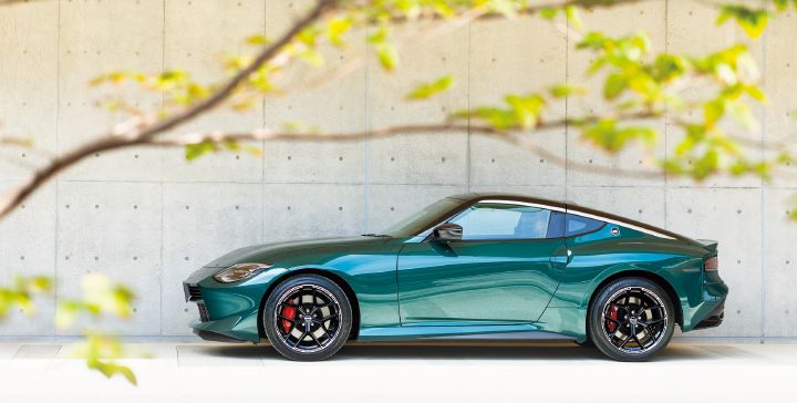 Unryu Green Fairlady Z photographed from the left-side profile in a bright outdoor setting, with the front of the vehicle facing toward the left side of the image. The car's long-hood, short-deck proportions, muscular rear fenders, black alloy wheels, and red brake calipers are clearly visible. A gray concrete wall forms the background, while softly blurred tree branches and green leaves frame the foreground. Natural light highlights the rich Unryu Green body color, emphasizing the refined styling and sculpted design of the Fairlady Z.