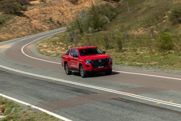 Elevated front three-quarter view of a 2026 Nissan Navara ST-X dual cab in Outback Red, driving along a winding sealed road through dry hillside terrain with scattered trees and scrub.