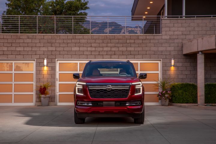 Front view of a red 2025 Nissan Armada parked in front of the garage attached to of a modern stone mansion at dusk.