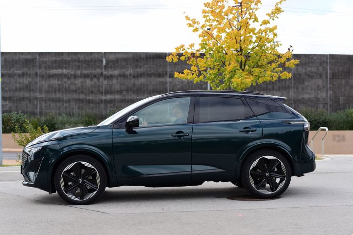 Seitenansicht eines dunkelgrünen Nissan Qashqai e‑Power mit Farer am Steuer, das Auto steht vor einer dunklen Wand und einem Baum mit gelbem Herbstlaub
