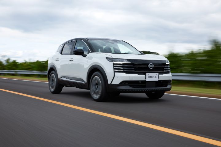 白系キックスの走行カット。車両は右向きに進み、斜め前方からの視点で撮影されている。背景には道路と緑の斜面、曇り空が広がる。