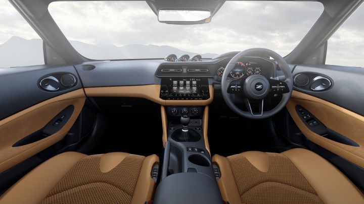 Interior view of a Fairlady Z from the rear seat perspective, facing forward toward the driver and passenger seats. The cabin features a two-tone design with camel brown seats and door panels contrasted by a black dashboard and center console. A touchscreen infotainment display is positioned in the center, with physical buttons and dials below it. The steering wheel, with a prominent “Z” logo, is visible along with analog gauges and digital displays behind it. Through the windows, a soft gray sky and faint mountain silhouettes can be seen, creating a calm and refined atmosphere.