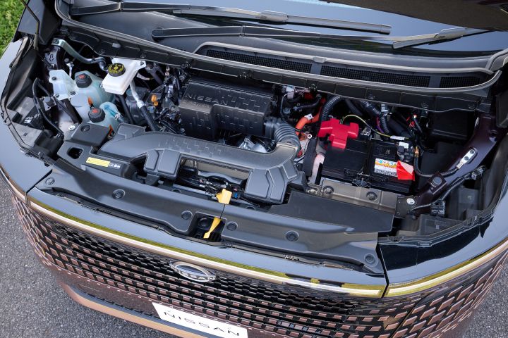 ボンネットを開けた状態の新型エルグランドのエンジンルームを、上方から見下ろすアングルで撮影した画像。中央には「NISSAN e‑POWER」と刻印されたブラックのエンジンカバーが配置され、その周囲に各種配管や電装部品が整理されている。右側には赤い端子カバー付きのバッテリー、左側には半透明の冷却液タンクや各種リザーバータンクが見える。下部にはフロントグリルの一部と「NISSAN」ロゴ入りナンバープレートが確認でき、全体は明るい屋外光の下で撮影されている。