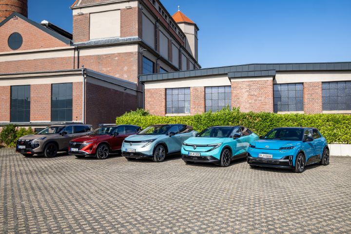 Vor einem Backsteingebäude und einer grünen Hecke stehen nebeneinander die fünf Nissan Modelle Nissan X‑Trail N-Trek, Nissan Qashqai, Nissan Ariya, Nissan Leaf und Nissan Micra, 3/4 Frontansicht