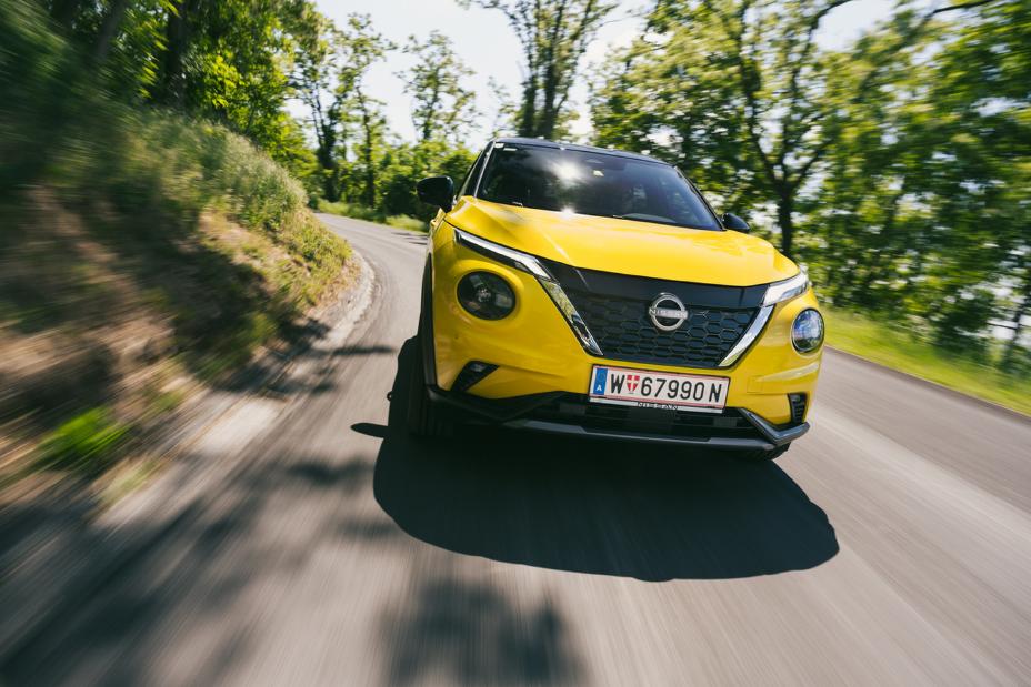 Dynamische Frontalaufnahme eines gelben Nissan Juke Hybrid, der um eine Landstraßenkurve fährt mit grünen Bäumen im Hintergrund