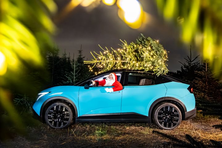 Der Weihnachtsmann lehnt sich aus dem Fahrerfenster eines türkisblauen Nissan LEAF, auf dem Dach transportiert er einen mit Lichterketten geschmückten Weihnachtsbaum