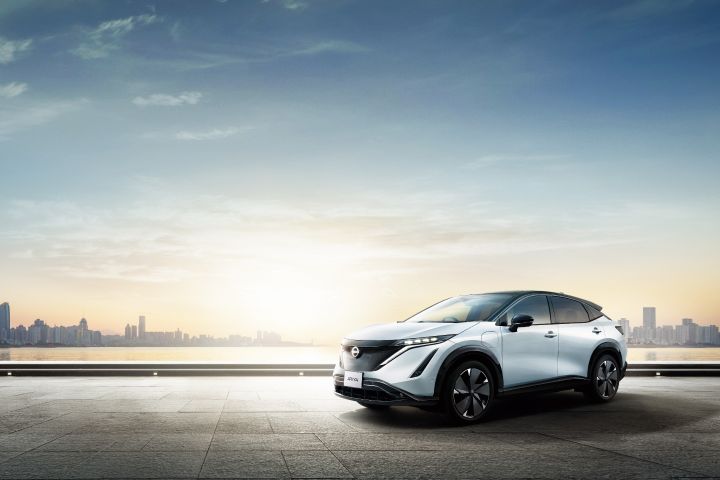 Ein weißer Nissan Ariya wird von der Morgensonne angestrahlt. Das Auto steht auf einem Platz, in der Ferne sind Gebäude sichtbar.