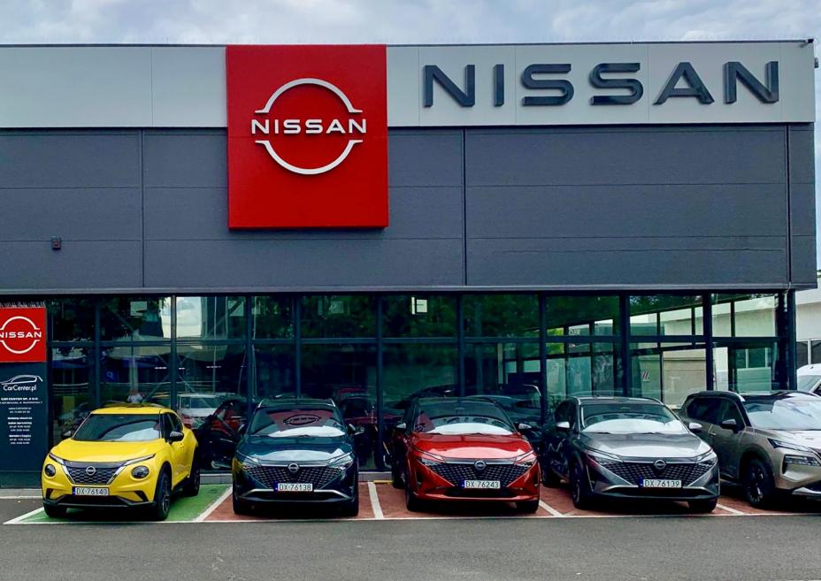 Front salonu Nissan z dużym czerwonym logo marki – pięć samochodów Nissan zaparkowanych przed wejściem, w tym żółty crossover, granatowy, czerwony i srebrny hatchback.