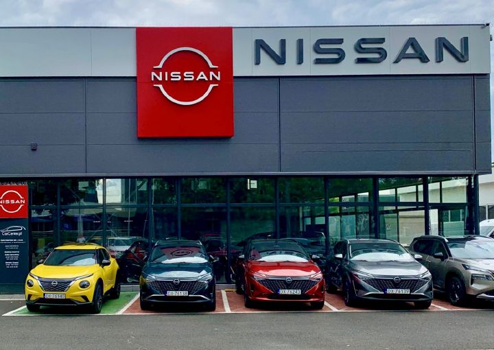 Front salonu Nissan z dużym czerwonym logo marki – pięć samochodów Nissan zaparkowanych przed wejściem, w tym żółty crossover, granatowy, czerwony i srebrny hatchback.