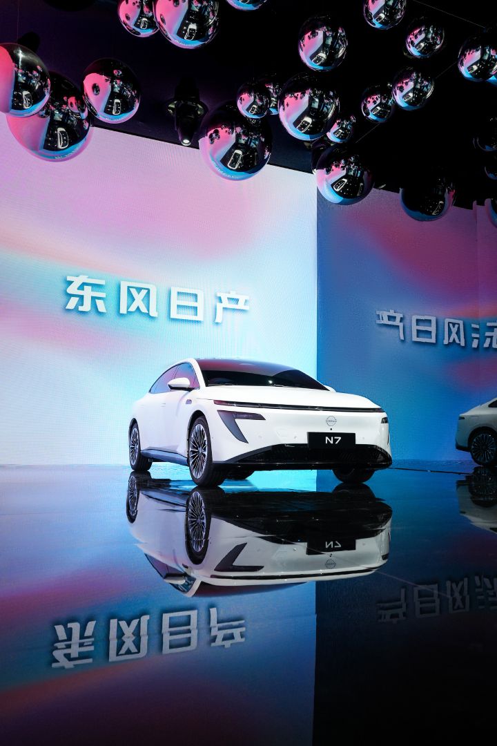 東風日産の白い新型セダン電気自動車「N7」の低位置からのショット。ピンクと青の背景、天井から銀色のボールが下がっている。