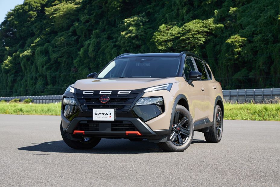 Front central image of a 2026 Nissan X‑TRAIL Rock Creek in a ‘Baja Storm’ with a black roof parked on the side of the road, shining in the sunlight with the backdrop of a green mountain and displaying on the orange accents and the tyres splayed out.
