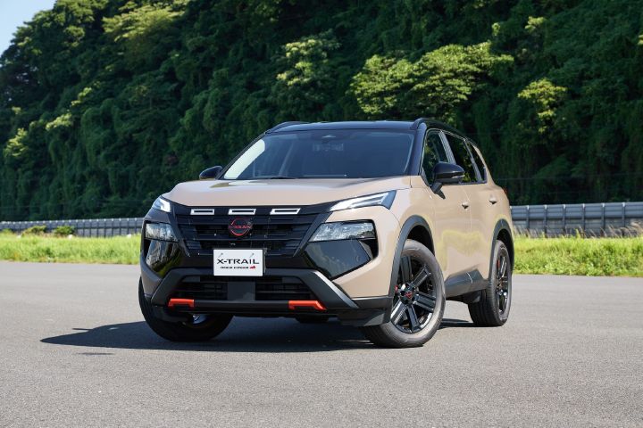 Front central image of a 2026 Nissan X‑TRAIL Rock Creek in a ‘Baja Storm’ with a black roof parked on the side of the road, shining in the sunlight with the backdrop of a green mountain and displaying on the orange accents and the tyres splayed out.