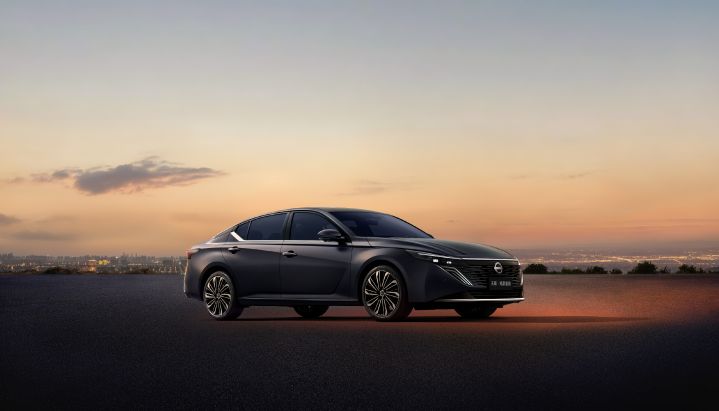 A black Teana parked on asphalt against a sunset sky. The car is facing left, showing its front and side. In the background, city lights stretch across the horizon, and the sky transitions from orange to blue.