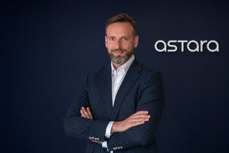 Mężczyzna w granatowym garniturze w prążki i białej koszuli stoi z założonymi rękami na tle granatowej ściany z białym logo Astara po prawej stronie.