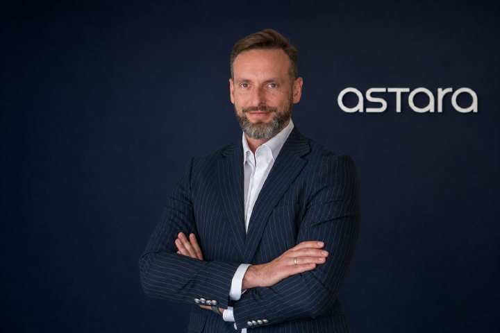 Mężczyzna w granatowym garniturze w prążki i białej koszuli stoi z założonymi rękami na tle granatowej ściany z białym logo Astara po prawej stronie.