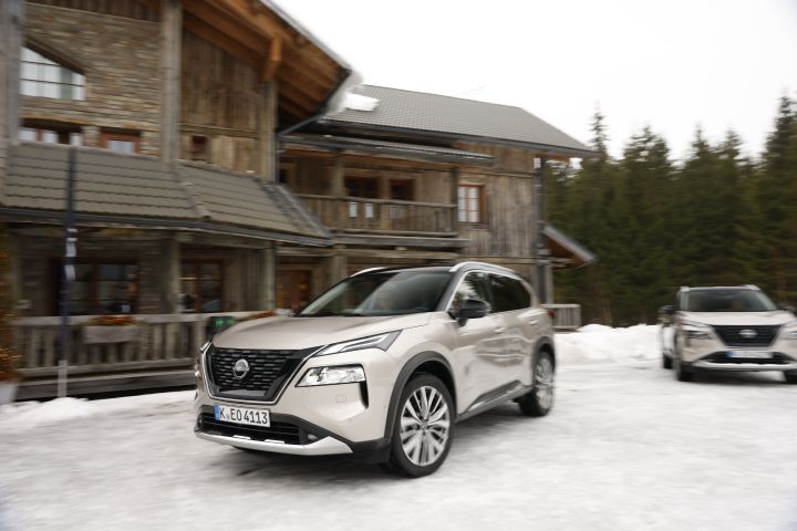 Dynamisches Bild von zwei silbernen Nissan X‑Trail e‑Power e‑4ORCE, die auf schneebedecktem Untergrund von einem Holzhaus wegfahren
