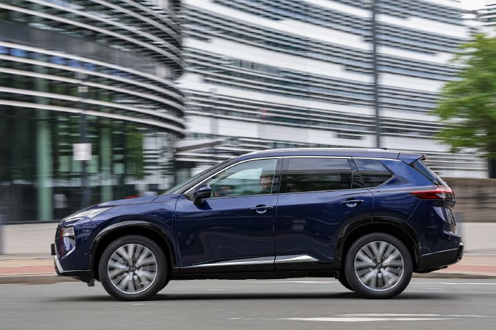 Seitliche Fahraufnahme eines dunkelblauen Nissan X‑Trail vor modernen Bürogebäuden