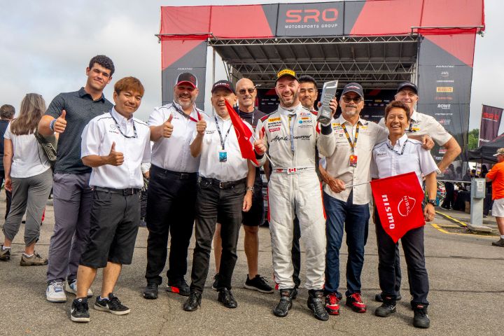 A group of men in various polos and racing passes stand by a racecar driver holding a trophy in front of a large pavilion structure.