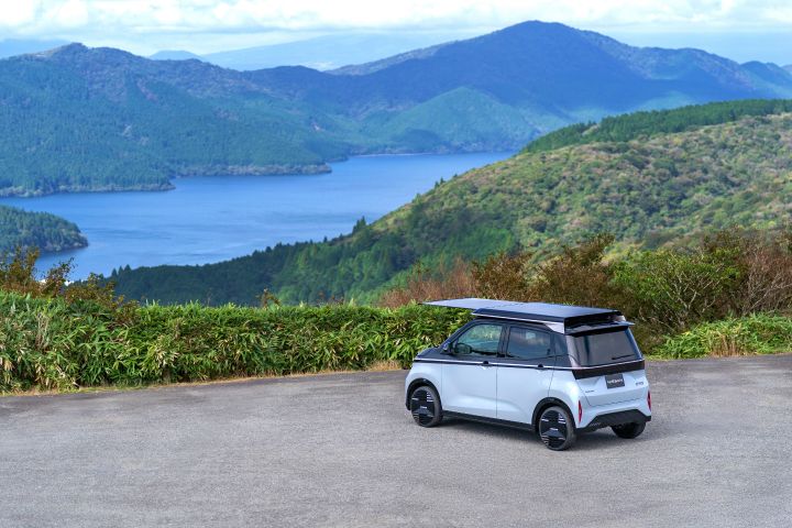 Against a backdrop of a lake and mountains, a light blue Nissan SAKURA is parked by the roadside, facing slightly left rear. The solar roof extends prominently forward, with green shrubs nearby and a blue lake and mountains in the distance.