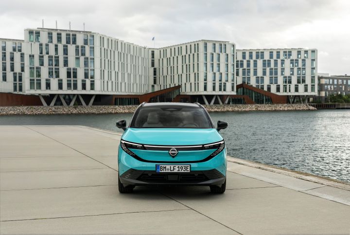 Frontalaufnahme eines türkisen Nissan LEAF, der am Meer steht, dahinter sieht man die helle Front eines großen modernen Gebäudes
