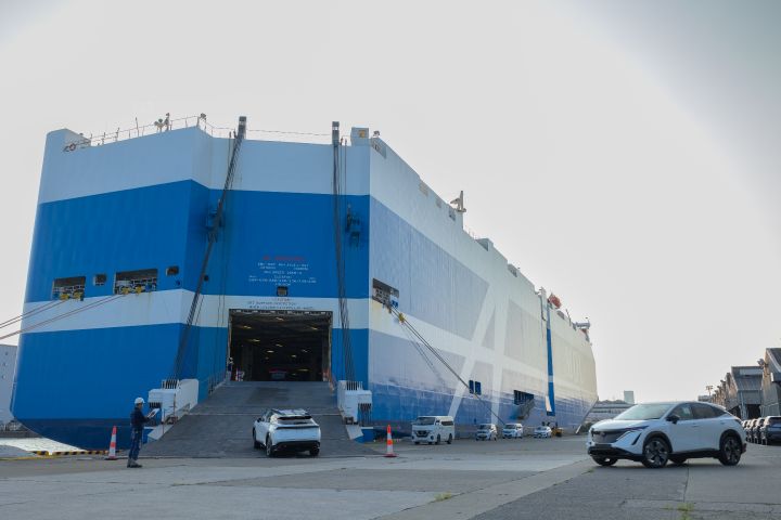 Image of two white Nissan Ariyas driving up from a docklands carpark into a heavy-duty container ship