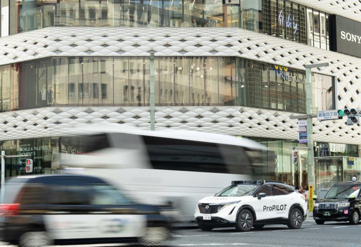 Ein weißer Nissan Ariya hält an einer Kreuzung in der Stadt, im Hintergrund ist das Gebäude Nissan Crossing zu sehen. Das Gebäude hat weiße Außenwände mit schwarzen Mustern und zahlreichen Glasfenstern. Weitere Fahrzeuge und Fußgänger sind ebenfalls zu sehen.