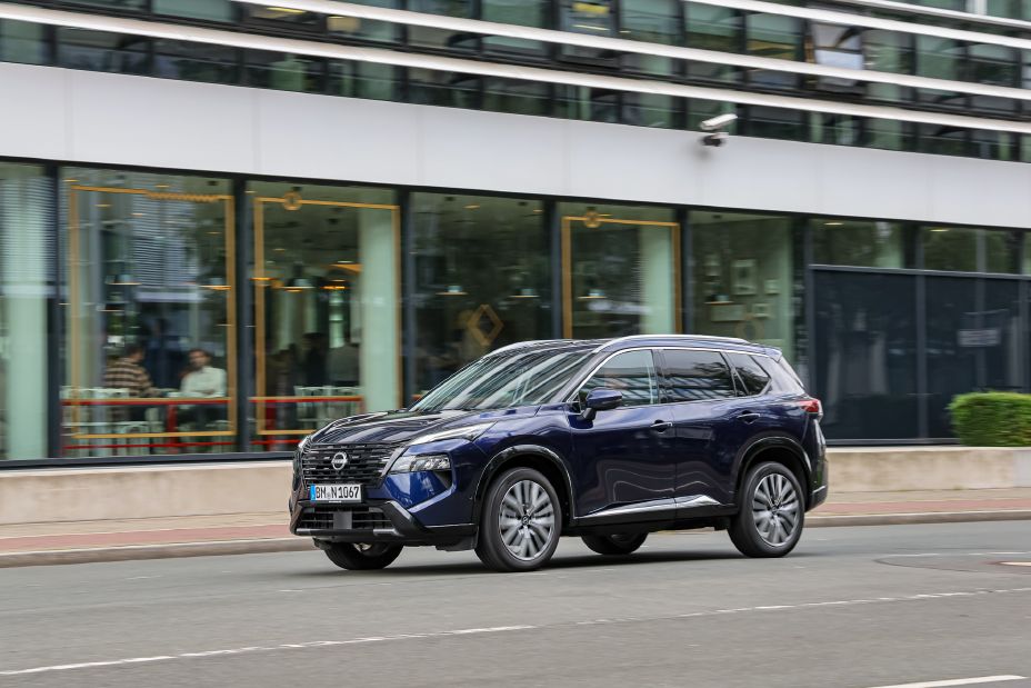Blick von schräg vorne auf einen dunkelblauen Nissan X‑Trail der an der gläsernen Front eines Cafés vorbeifährt