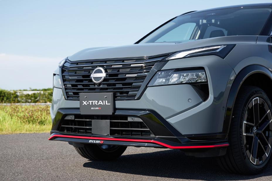 A grey Nissan X-Trail NISMO is parked on a road. The vehicle is captured from the front, featuring a prominent grille with the Nissan logo, sleek headlights, and red accents on the lower bumper. The background includes greenery and a clear sky.