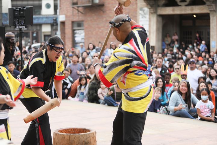 Two men dressed in yellow, black, and red traditional attire swing wood hammers over a bucket in front of a crowd.