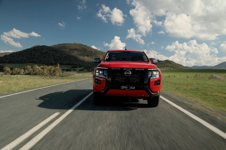 Aerial view of a 2026 Nissan Navara ST‑X Outback Red dual cab driving along a sealed road positioned beside a steep hillside covered with grass and scattered trees.