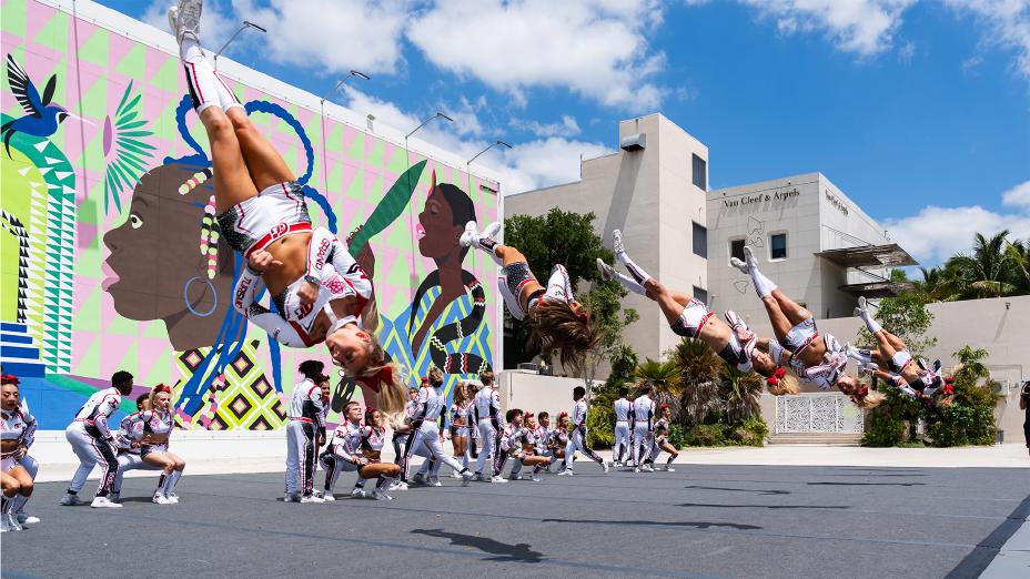 Atletas da equipe de cheerleading “Sakura Storm” realizam uma sequência de mortais no ar durante uma performance estilo flash mob na Jungle Plaza, em Miami. Os integrantes usam uniformes temáticos nas cores branco, vermelho e preto com detalhes que remetem ao automobilismo, enquanto o mural colorido ao fundo celebra a cultura local. A ação faz parte da parceria com a Equipe Nissan de Fórmula E para o E-Prix de Miami.