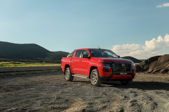 Wide front three-quarter view of a 2026 Nissan Navara ST-X dual cab in Outback Red, stationary on a broad gravel surface with hills, pale sky, and mounded earth in the distance.