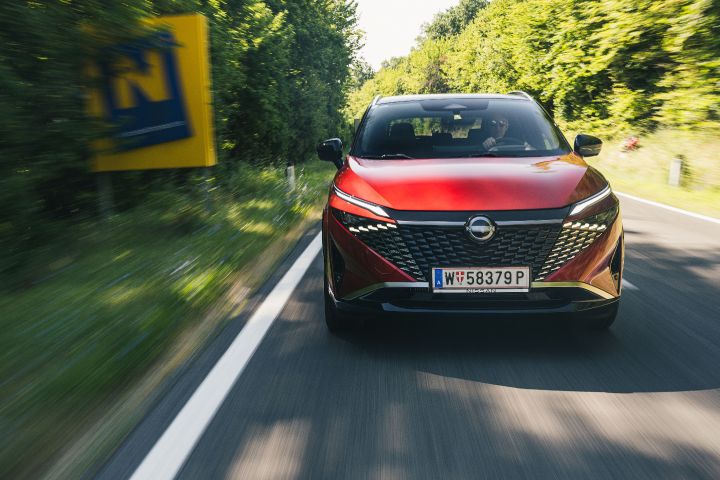 Frontale Fahraufnahme eines Nissan Qashqai in Fuji Sunset Red auf einer Landstraße mit Bäumen und einem gelben Schild am Fahrbahnrand