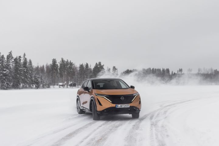 Elektroauto Nissan Ariya e‑4ORCE in Akatsuki Copper mit Dach und Außenspiegel in Pearl Black driftet auf Eissee mit schneebedeckten Bäumen im Hintergrund, 3/4 Front