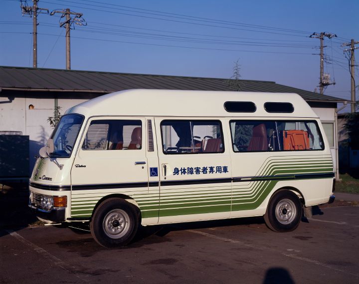 A white and green retro van with Japanese text on the side is parked on a paved road. The van has large windows and a roof extension