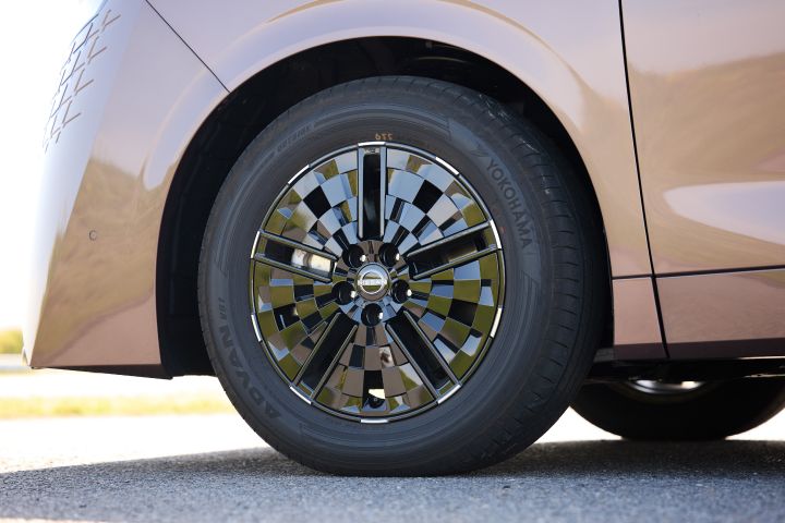 新型エルグランドのフロントホイールをクローズアップした画像で、側面からの視点で捉えられている。ブラックの多スポークホイールには幾何学的な光沢デザインが施され、中央には「NISSAN」ロゴが配置されている。タイヤ側面には「YOKOHAMA」の文字が見え、ホイール周囲のボディは滑らかな曲面でブロンズカラーに輝いている。上部にはホイールアーチとフェンダーがあり、下部には灰色の舗装路面が広がる。背景は明るくぼかされ、日中の屋外環境が感じられる。