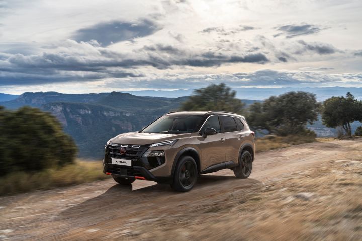 Der neue 2026 Nissan X‑Trail N-Trek in der Karosseriefarbe „Baja Storm“ steht auf einem Feldweg mit Blick auf ein großes Gewässer, das von bewaldeten Hügeln umgeben ist. Die Szene unterstreicht das Outdoor-Abenteuer und die Geländegängigkeit mit natürlicher Vegetation und ruhigem Wasser im Hintergrund.