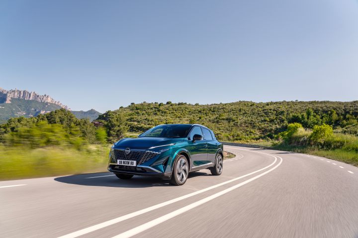 Ein dunkelgrün-blauer Nissan Qashqai mit e‑Power-Antrieb der nächsten Generation fährt bei Sonnenschein durch eine langgestreckte Rechtskurve, im Hintergrund ein bewaldeter Hügel und blauer Himmel