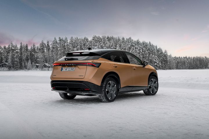 Elektroauto Nissan Ariya e‑4ORCE in Akatsuki Copper mit Dach und Außenspiegel in Pearl Black auf Eissee mit schneebedeckten Bäumen im Hintergrund, 3/4 Heck