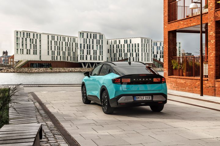 Foto von schräg hinten von einem türkisen Nissan LEAF mit schwarz abgesetzten Heckfenster, rechts davon ein rotes Backsteingebäude und im Hintergrund ein weißes Gebäude