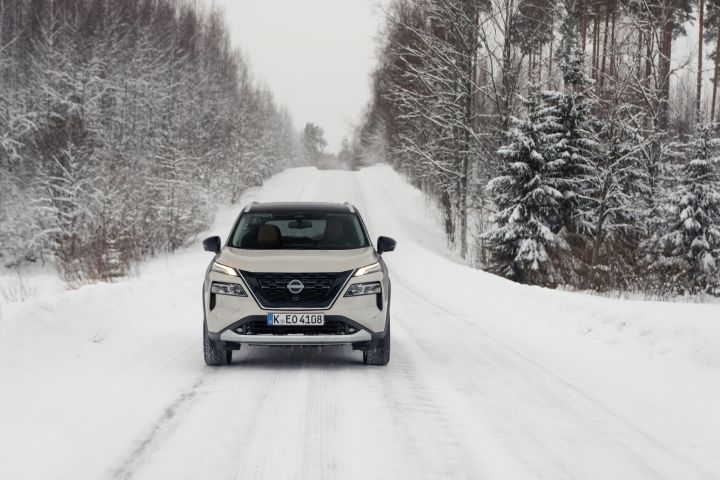 Silberner Nissan X‑Trail e‑Power e‑4ORcE fährt auf schneebedeckter Straße mit verschneiten Bäumen auf beiden Fahrbahnseiten, Front