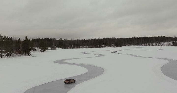 Elektroauto Nissan Ariya e‑4ORCE in Akatsuki Copper mit Dach und Außenspiegel in Pearl Black fährt auf Eissee mit schneebedeckten Bäumen im Hintergrund, kleines Auto in weißer Landschaft aus der Drohnen-Perspektive